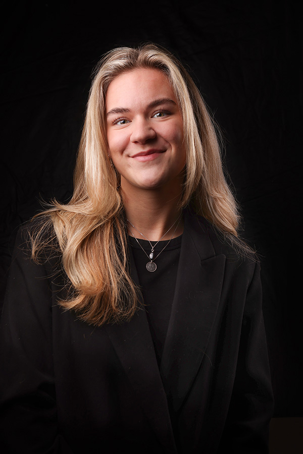 een blonde jongevrouw met lichte ogen kijkt recht in de camera. de verlichting is alleen op haar gezicht. ze is zwart gekleed en de achtergrond is tevens zwart. gefotografeerd in de studio in haarlem